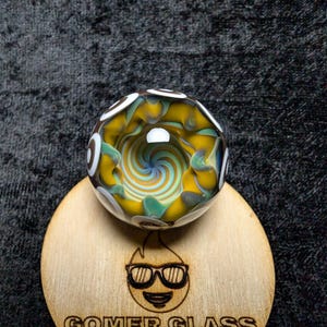 Könnte beinhalten: Eine farbenfrohe Glaskugel mit gelben, türkisfarbenen und blauen Wirbeln, die auf einem Holzuntersetzer liegt. Die Murmel hat weiße und schwarze Akzente. Der Untersetzer trägt den Text "GOMER GLASS" und ein Logo einer lächelnden Flamme mit Sonnenbrille.