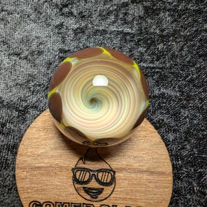 May include: A brown and yellow glass marble with a swirling white and blue pattern inside. The marble is sitting on a wooden coaster with the words "Gomer Glass" and a cartoon image of a person wearing sunglasses.