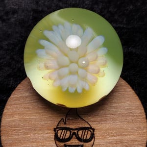 May include: A glass marble with a white flower design inside. The marble is sitting on a wooden coaster with the logo "Gomer Glass" and a cartoon face with sunglasses.