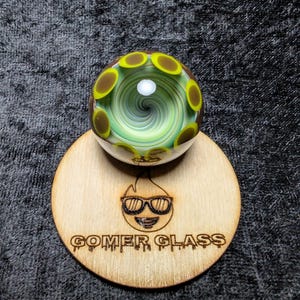 May include: A glass marble with a swirling green and white interior, accented by brown and yellow circular designs. It rests on a wooden stand with the text "GOMER GLASS" and a smiling flame logo.