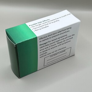 Op de afbeelding: Groene doos met witte tekst die de bijwerkingen en dosering van een medicijn beschrijft. De tekst bevat de zin "asshole-ness can be infectious." De doos heeft een lege ruimte voor de naam van de pati&euml;nt.