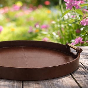 May include: A brown, oval serving tray with stitched detailing and wooden handles. The tray is made of a leather-like material and sits on a weathered wooden surface. The background features a blurred garden with pink flowers.