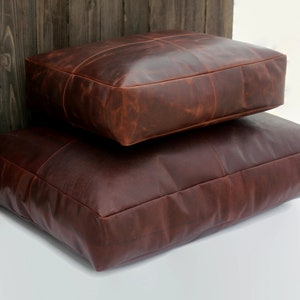 May include: Two rectangular, brown leather floor cushions stacked on a white surface. The cushions have a stitched, grid-like pattern and a rich, reddish-brown colour. The cushions are placed next to a wooden wall.