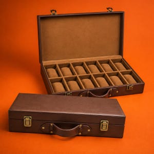 Puede incluir: Un estuche rectangular de color marrón oscuro para guardar relojes, con asa y dos cierres metálicos de color latón. El estuche abierto muestra doce compartimentos individuales, cada uno con un cojín marrón suave para sujetar relojes.