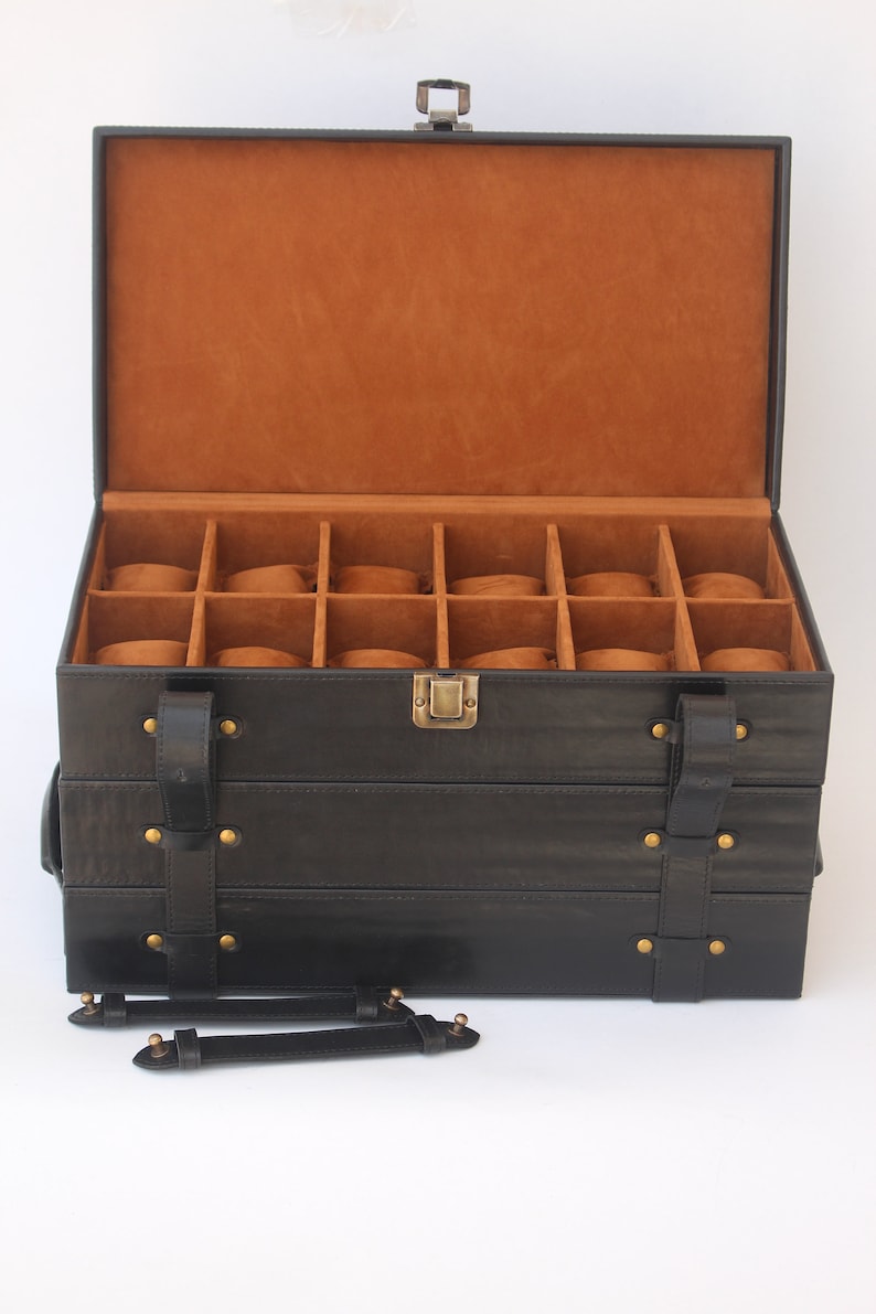 Puede incluir: Una caja de almacenamiento para relojes rectangular de color negro con tapa abatible e interior de color tostado. La caja cuenta con doce compartimentos individuales, cada uno con una almohadilla. El exterior tiene correas de cuero negro.