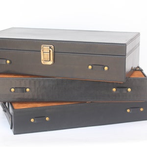 Puede incluir: Una pila de tres cajas de almacenamiento rectangulares con lados exteriores negros y una superficie superior de color madera natural. Cada caja cuenta con peque&ntilde;as asas laterales de aspecto cuero negro aseguradas con remaches met&aacute;licos dorados.