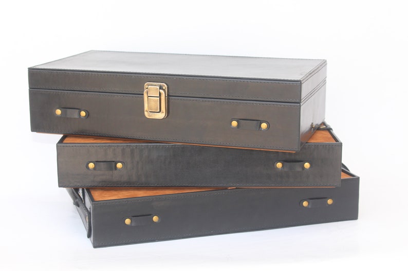 Puede incluir: Una pila de tres cajas de almacenamiento rectangulares con lados exteriores negros y una superficie superior de color madera natural. Cada caja cuenta con peque&ntilde;as asas laterales de aspecto cuero negro aseguradas con remaches met&aacute;licos dorados.