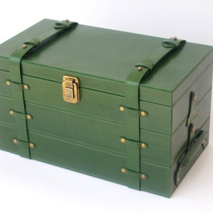 Puede incluir: Una caja de almacenamiento rectangular de color verde bosque con acabado liso, que presenta dos correas verticales verdes aseguradas con remaches de metal en tono lat&oacute;n. La caja incluye un cierre de pestillo central de metal y un asa lateral.