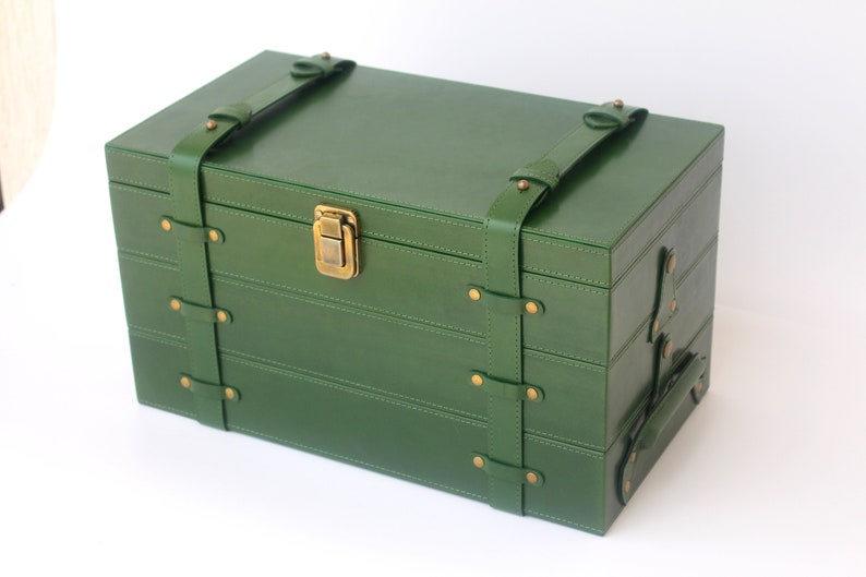 Puede incluir: Una caja de almacenamiento rectangular de color verde bosque con acabado liso, que presenta dos correas verticales verdes aseguradas con remaches de metal en tono lat&oacute;n. La caja incluye un cierre de pestillo central de metal y un asa lateral.