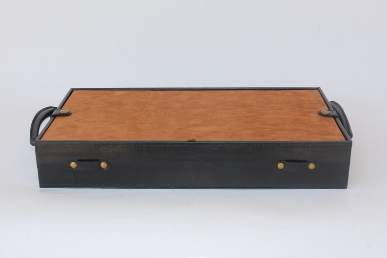 Puede incluir: Una bandeja rectangular con un marco exterior negro y una base interior de color tostado. La bandeja incluye dos asas negras en los laterales, cada una asegurada con remaches met&aacute;licos de color lat&oacute;n, y una superficie plana y lisa.