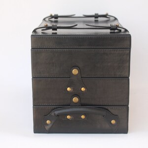 Puede incluir: Un estuche de almacenamiento rectangular negro con tres compartimentos apilados. El estuche presenta tachuelas met&aacute;licas de color lat&oacute;n, correas con hebillas en la parte superior y un asa resistente en la secci&oacute;n inferior frontal, asegurado con herrajes met&aacute;licos.