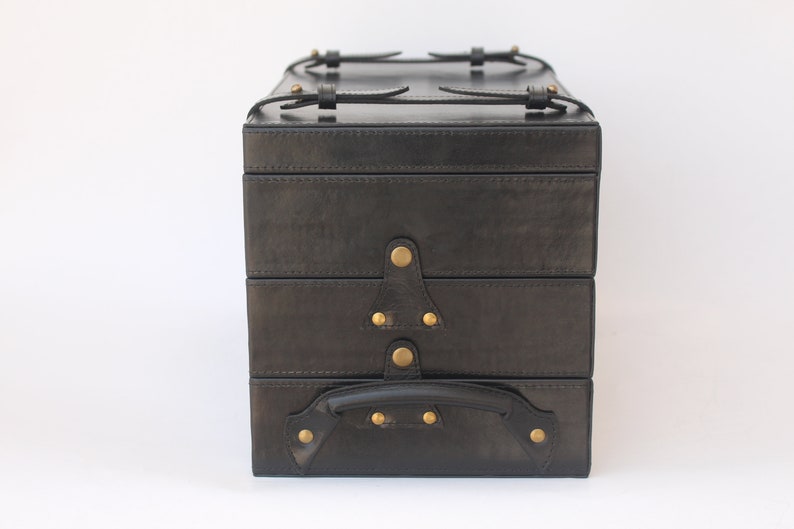 Puede incluir: Un estuche de almacenamiento rectangular negro con tres compartimentos apilados. El estuche presenta tachuelas met&aacute;licas de color lat&oacute;n, correas con hebillas en la parte superior y un asa resistente en la secci&oacute;n inferior frontal, asegurado con herrajes met&aacute;licos.