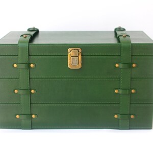 Puede incluir: Una caja de almacenamiento rectangular de color verde oscuro con un acabado liso. La caja cuenta con dos correas verticales verdes aseguradas con remaches de metal color lat&oacute;n y un cierre central de metal color lat&oacute;n en la tapa frontal.