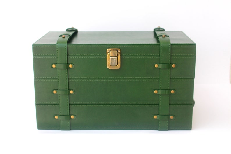 Puede incluir: Una caja de almacenamiento rectangular de color verde oscuro con un acabado liso. La caja cuenta con dos correas verticales verdes aseguradas con remaches de metal color lat&oacute;n y un cierre central de metal color lat&oacute;n en la tapa frontal.