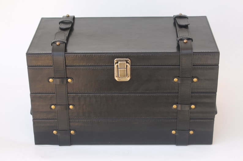 Puede incluir: Un cofre de almacenamiento rectangular negro con dos correas verticales de aspecto cuero con remaches y hebillas de metal en tono lat&oacute;n. La parte frontal del cofre incluye un mecanismo de cierre central de metal en tono lat&oacute;n.