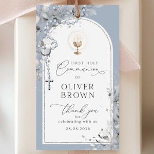 Puede incluir: Una caja de regalo marfil con una cinta melocotón y una etiqueta. La etiqueta, de color azul claro, presenta detalles florales y el texto "First Holy Communion of Oliver Brown". Un pequeño broche de ángel en la parte inferior.