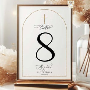 May include: A gold-framed table number card with the number 8 in black script. The card includes the words "Table" and "Baptism" with the name "Oliver Brown" and the date 08.08.2020. The card is displayed on a gold stand.