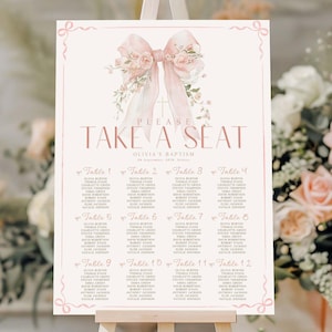 May include: A light pink seating chart with a floral bow design and the text "PLEASE TAKE A SEAT". The chart lists names under table numbers 1-12. The event is for Olivia's Baptism.
