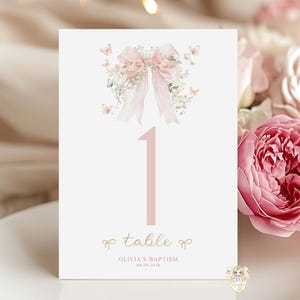 Può includere: Un biglietto bianco con numero di tavolo, con fiocco rosa e design floreale. Il numero "1" è rosa, con la parola "table" e il nome "Olivia's Baptism" stampati sotto. Il biglietto è su un piatto bianco, con fiori sullo sfondo.