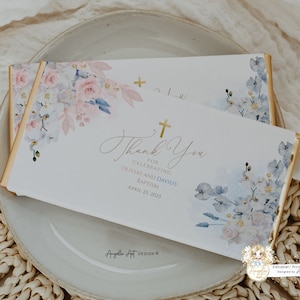 May include: A white chocolate bar wrapper with a floral design in shades of pink and blue. The wrapper features a gold cross and the text "Thank You for celebrating Olivia's and David's Baptism April 25, 2025".