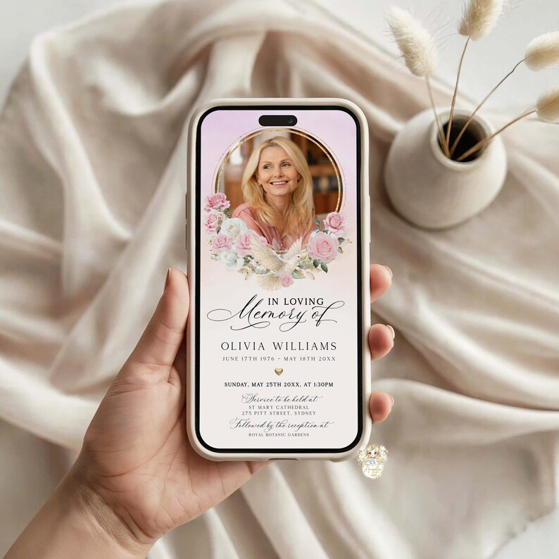 May include: A smartphone displaying a memorial announcement. The screen features a photo of a woman in a gold frame, surrounded by pink and white flowers. Text reads "In Loving Memory of Olivia Williams" with dates and service details.