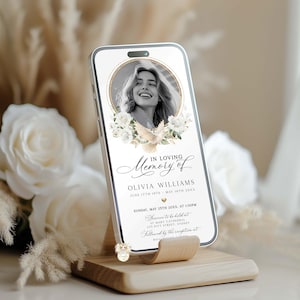 May include: A smartphone displays a memorial design with a photo in a gold frame, white flowers, and a dove. Text reads "In Loving Memory of Olivia Williams" with dates and service details. The phone rests on a wooden stand.