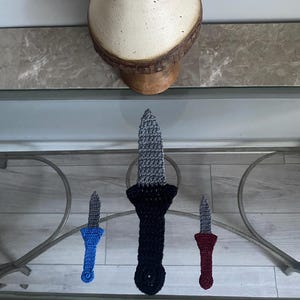 Könnte beinhalten: Drei gehäkelte Messer, eines blau, eines schwarz und eines rot, werden auf einer Glasfläche ausgestellt.