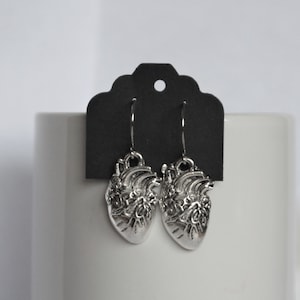 Puede incluir: Un par de pendientes de corazón colgantes de plata con detalles intrincados. Los pendientes cuelgan de una tarjeta negra con borde festoneado.