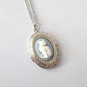 May include: A silver chain necklace with a silver locket featuring a white porcelain cat on a light blue background.
