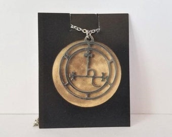 May include: A silver chain necklace with a round pendant featuring a black and gold sigil. The sigil is a circle with a cross inside and a letter L in the center.