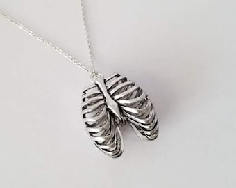 Puede incluir: Un collar de plata con un colgante de caja torácica. El colgante es detallado y realista, con una cadena delicada.