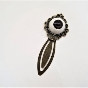 May include: A bronze-colored metal bookmark with an eye-shaped design. The eye has a white iris and a dark pupil, set within an ornate, circular frame. The bookmark's stem is elongated and oval.