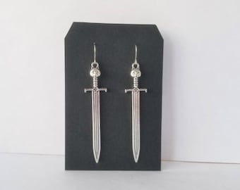 May include: A pair of silver sword earrings. The swords have a detailed design and are hanging from a black display card.