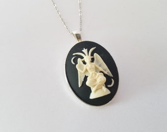 May include: A silver necklace featuring an oval pendant with a black background. The pendant showcases a cream-colored relief of a winged figure. The necklace is displayed against a white background.