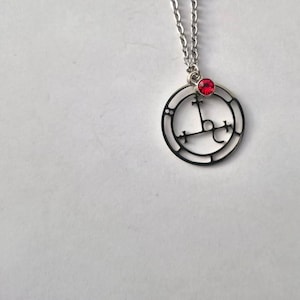 May include: A silver necklace with a circular pendant featuring a black sigil and a red gemstone.
