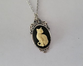 Può includere: Una collana con catena d'argento con un ciondolo a cammeo che presenta un gatto bianco seduto su uno sfondo nero. Il cammeo è circondato da una cornice ornata d'argento.