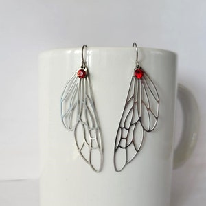 May include: A pair of silver earrings with a delicate, intricate wing design and a red crystal accent.