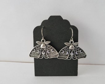 May include: A pair of silver earrings featuring a moth design with moon phases. The earrings are hanging from a black display card.