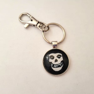 Könnte beinhalten: Ein runder Schlüsselanhänger mit einem schwarzen Hintergrund und einer weißen Totenkopf-Grafik. Der Schlüsselanhänger hat einen silberfarbenen Metallring und einen Karabinerhaken zum Befestigen an einer Tasche oder Schlüsseln. Das Totenkopf-Design ist ein bekanntes Symbol.