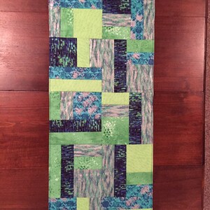 Puede incluir: Un camino de mesa de patchwork verde y azul con un patr&oacute;n geom&eacute;trico. El camino de mesa est&aacute; hecho de cuadrados de tela cosidos juntos.