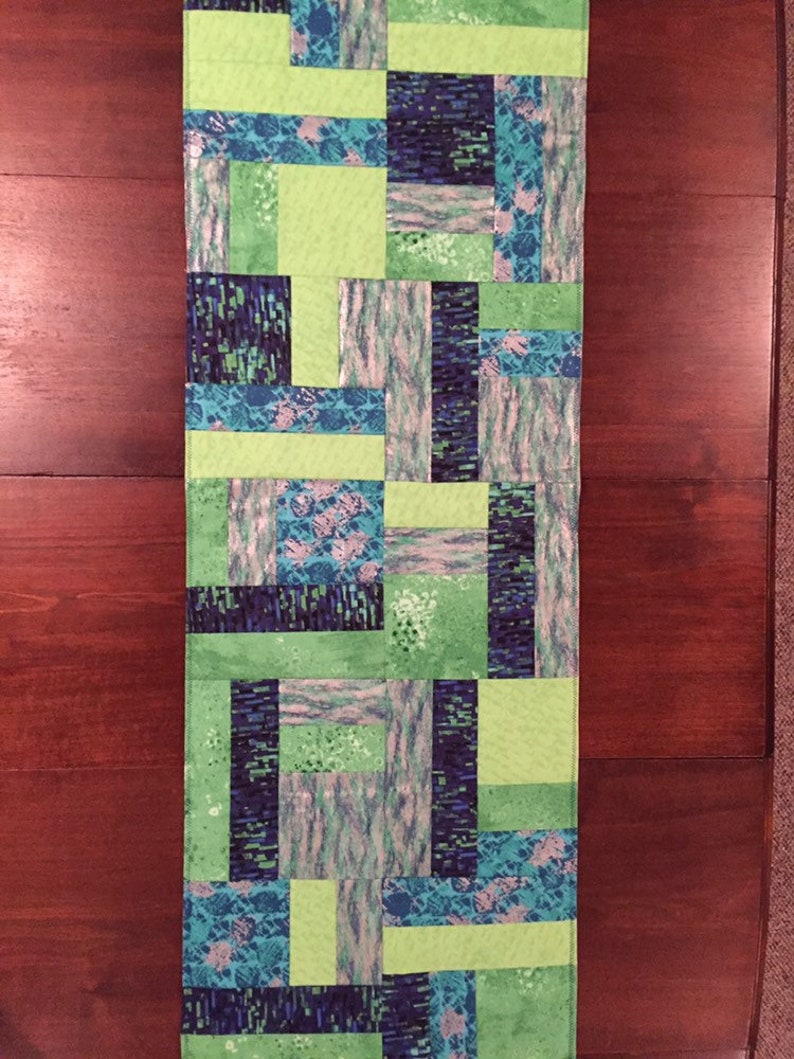 Puede incluir: Un camino de mesa de patchwork verde y azul con un patr&oacute;n geom&eacute;trico. El camino de mesa est&aacute; hecho de cuadrados de tela cosidos juntos.