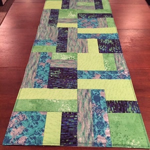 Puede incluir: Un camino de mesa de patchwork verde y azul con un patr&oacute;n geom&eacute;trico. El camino de mesa est&aacute; hecho de diferentes telas con varias texturas y colores.