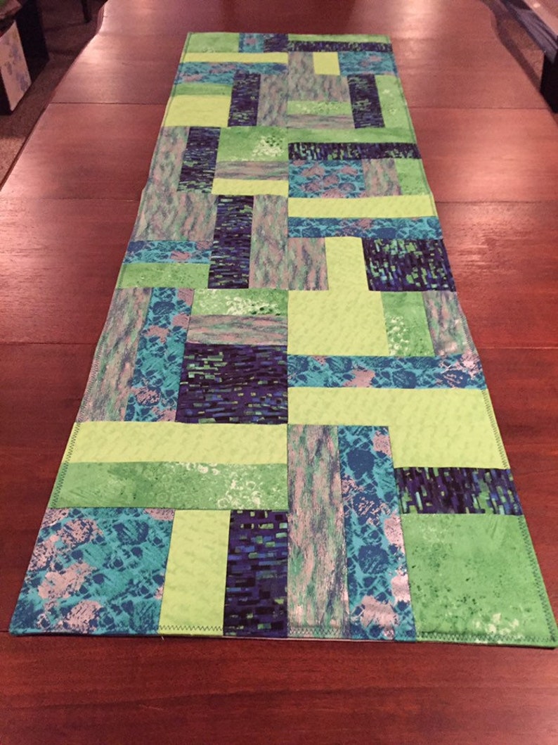 Puede incluir: Un camino de mesa de patchwork verde y azul con un patr&oacute;n geom&eacute;trico. El camino de mesa est&aacute; hecho de diferentes telas con varias texturas y colores.