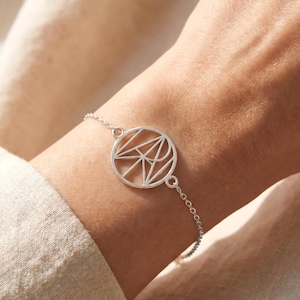 Op de afbeelding: Een zilverkleurige armband met een ronde hanger voorzien van een geometrisch, opengewerkt ontwerp van kruisende lijnen. De hanger is bevestigd aan een fijne schakelketting die om de pols valt, wat zorgt voor een strakke, metalen uitstraling.