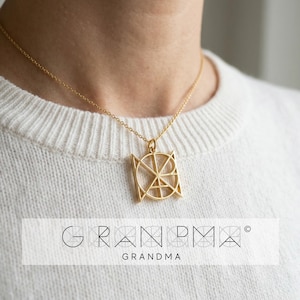 Puede incluir: Un collar con colgante dorado con la palabra "GRANDMA" en un diseño geométrico. El colgante cuelga de una delicada cadena. La palabra "GRANDMA" también está impresa en una fuente estilizada debajo.