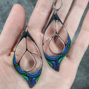 May include: A pair of dangle earrings with a black, green, and purple design. The earrings are made of polymer clay and have a clear, glass-like center.