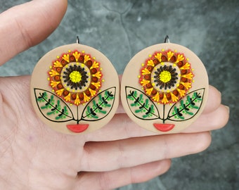 Op de afbeelding: Een paar ronde, beige oorbellen met een kleurrijk bloemenontwerp. De bloemen hebben gele centra, oranje bloemblaadjes en groene bladeren. De oorbellen hebben een rode lipvorm aan de onderkant.