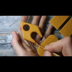 May include: A hand holding a paintbrush and painting a yellow, textured, geometric shape on a clear glass surface.
