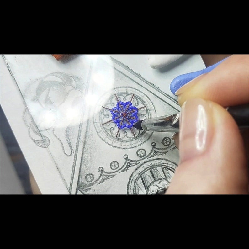 May include: A close-up of a hand painting a blue and brown mandala design on a white surface. The design is surrounded by a black and gray intricate border.