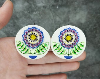Op de afbeelding: Een paar witte keramische oorbellen met een blauw, geel, groen en paars bloemenontwerp.